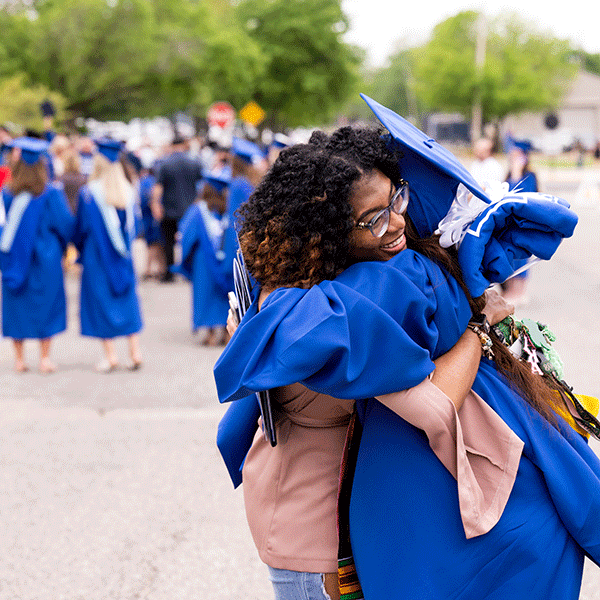 A smiling young woman of color with glasses and dark, curly hair hugs an Indiana State graduate in blue cap and gown outdoors on a spring day. Other graduates and guests are visible in the background, as are many green trees, traffic signs, and buildings in the distance.